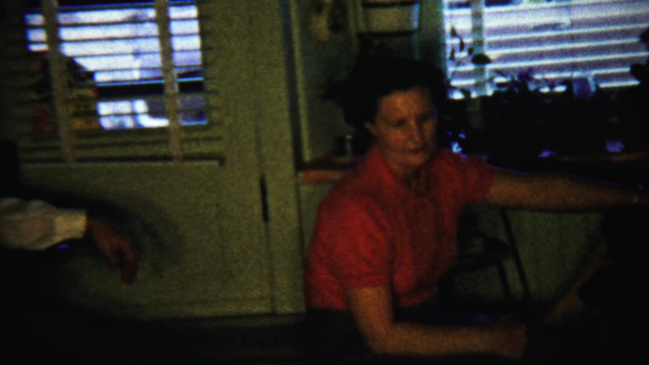 A woman in a red blouse looks toward the camera inside a dim 1958 reunion setting in Saint Francis, Kansas.