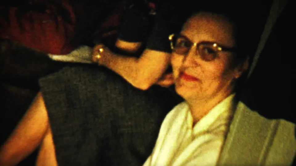 A woman in glasses and a light blouse faces the camera while other adults sit nearby in the 1958 Saint Francis reunion footage.
