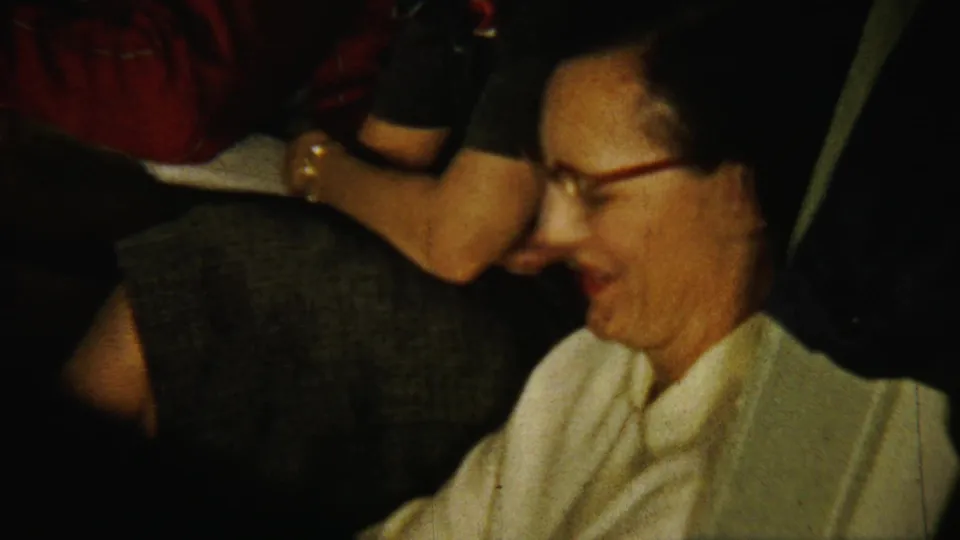 A woman in glasses smiles in profile beside another seated adult during the 1958 Saint Francis, Kansas indoor gathering.