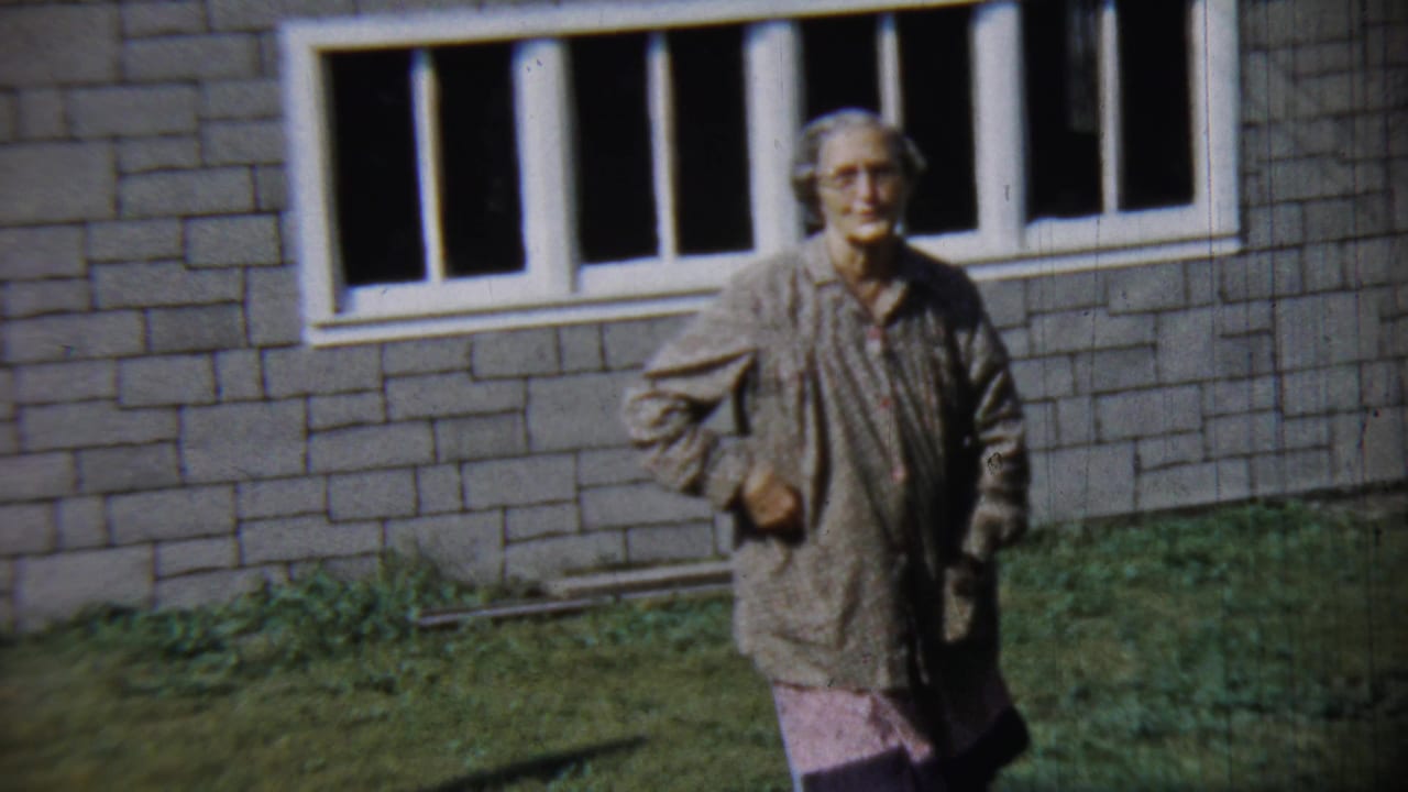 Older woman walking on grass in front of a brick house with white windows.