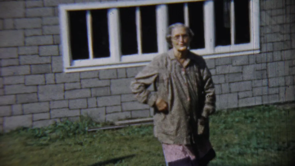 Older woman in glasses and a patterned housecoat walks across a lawn in Chadron, Nebraska, in 1959.