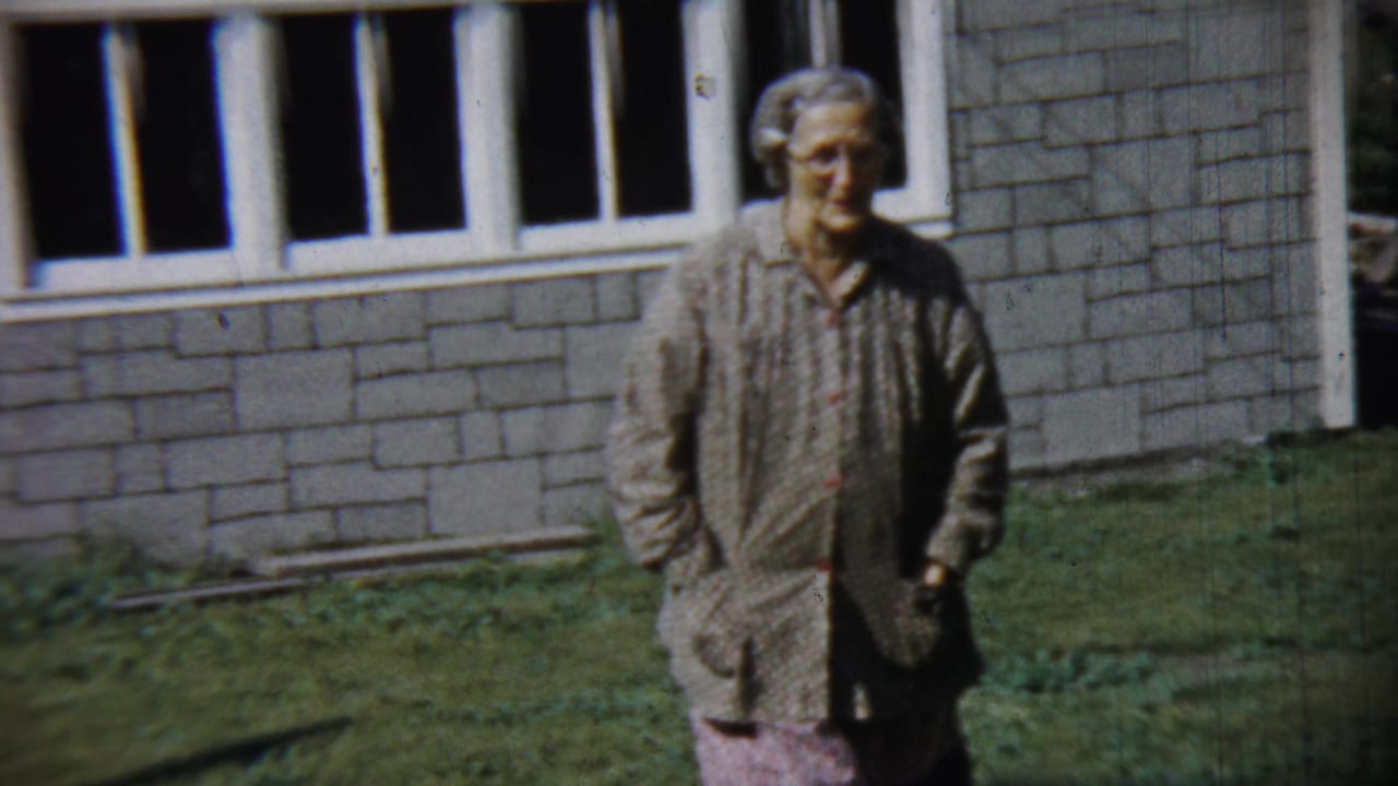 Older woman standing or walking on a lawn beside a brick house wall.