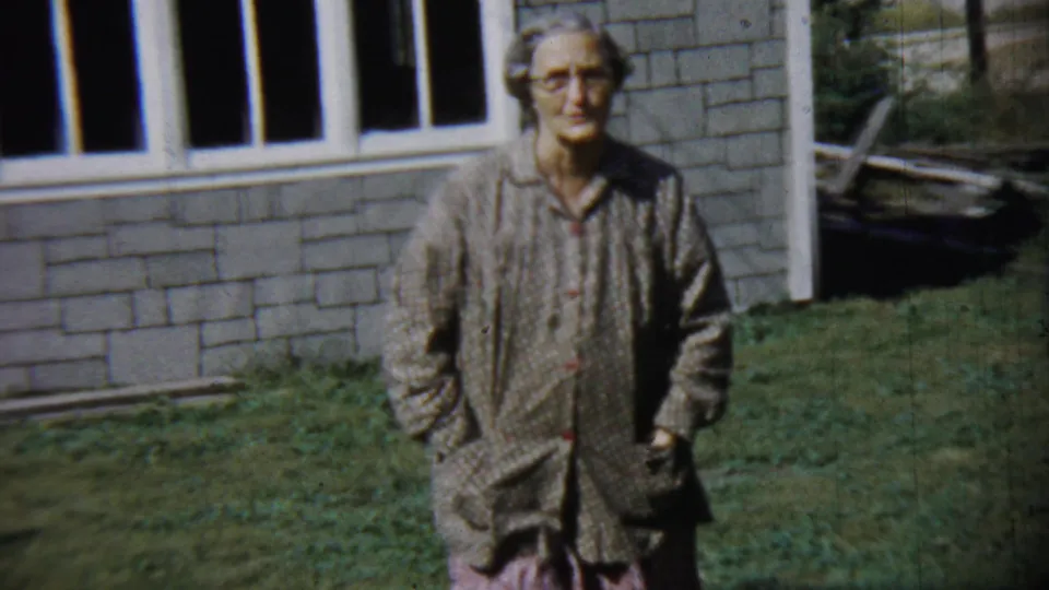 In this Chadron, Nebraska frame, the older woman walks nearer the camera against the brick house and lawn.