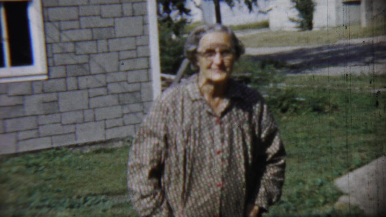 Elderly woman in glasses stands smiling in a grassy yard beside a stone house.