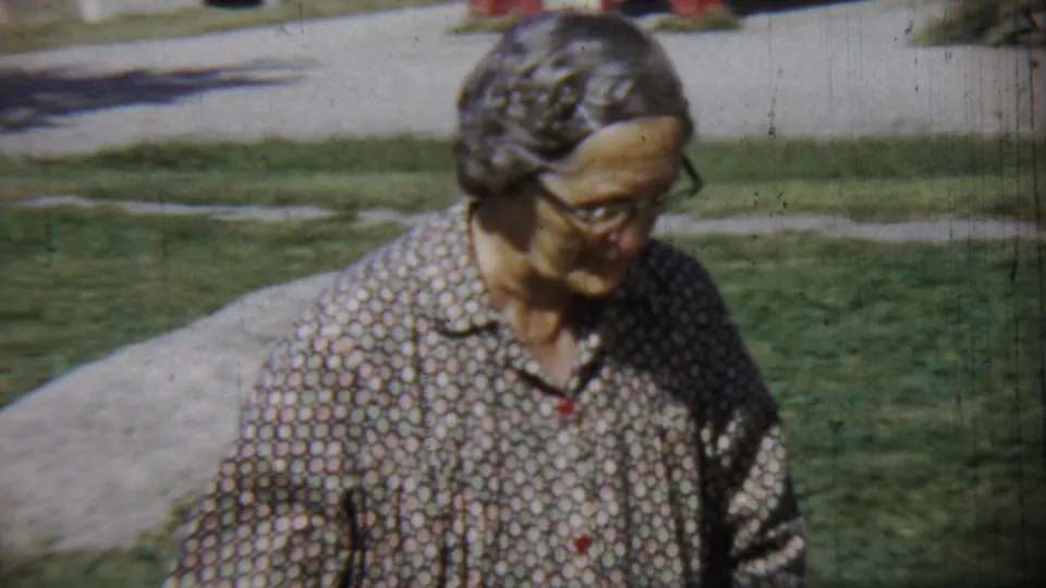 A closer frame shows the elderly woman in glasses and a patterned shirt moving across the Chadron yard in bright daylight.