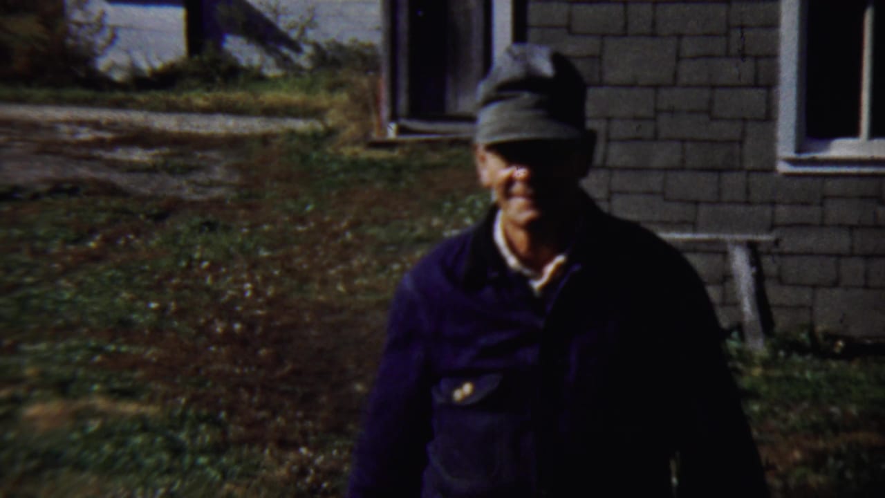 Man in a grey cap and dark jacket walking toward the camera outside a house.
