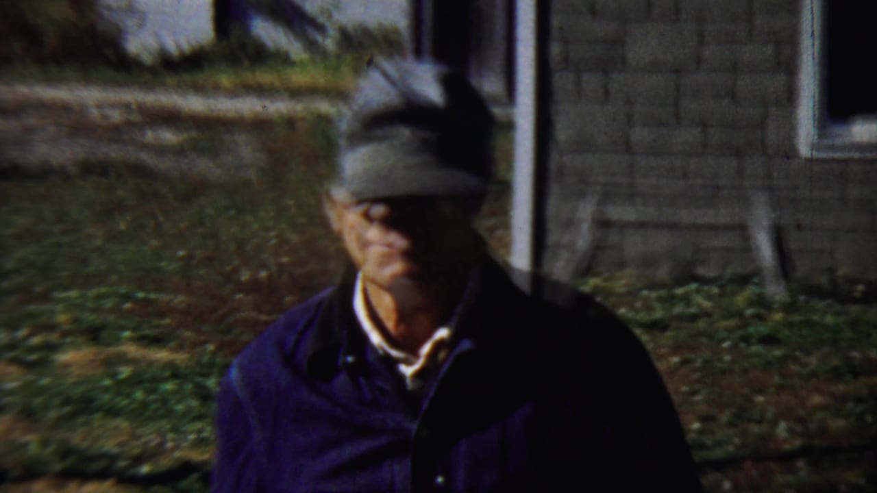 Man in a grey cap closer to the camera with a house and yard behind him.