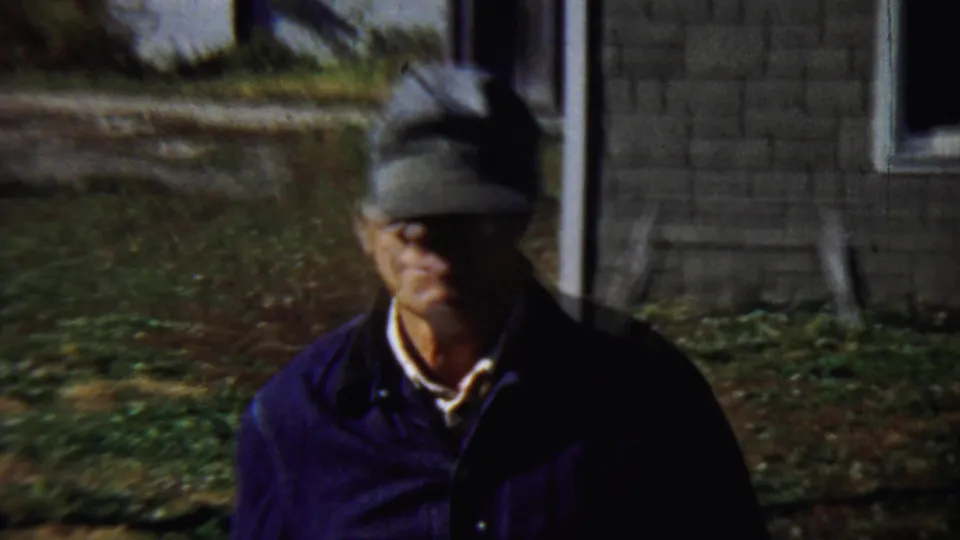 The man in a grey cap and dark jacket appears closer to the camera, with residential windows and a yard behind him.