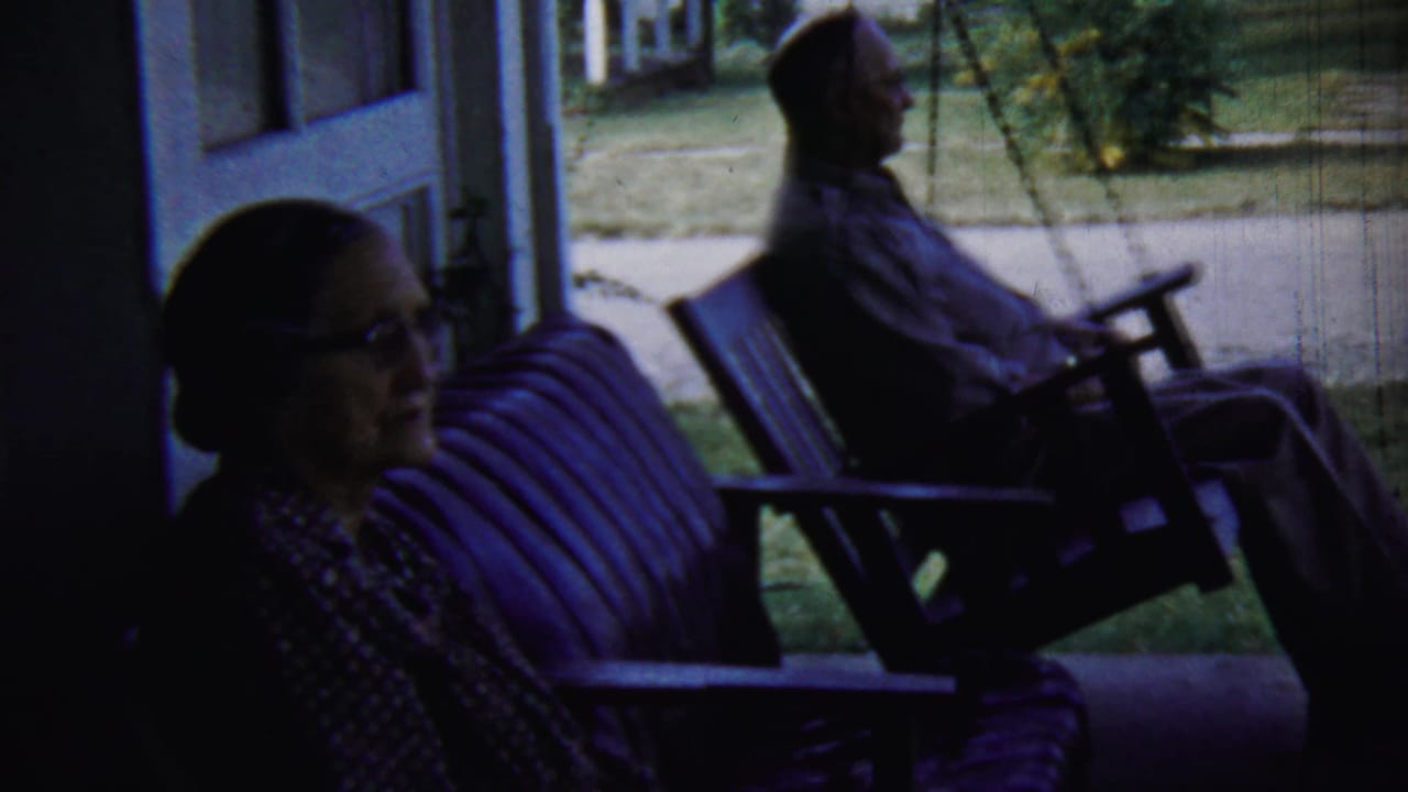 Two adults sit on a shaded porch swing with a lawn and sidewalk beyond.