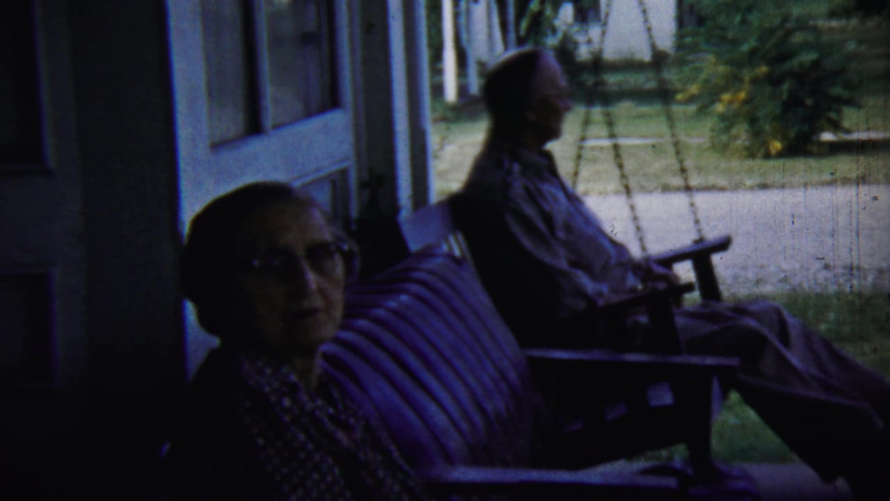 The woman turns toward the camera on a shaded Chadron porch while the man remains seated in profile on the swing behind her.