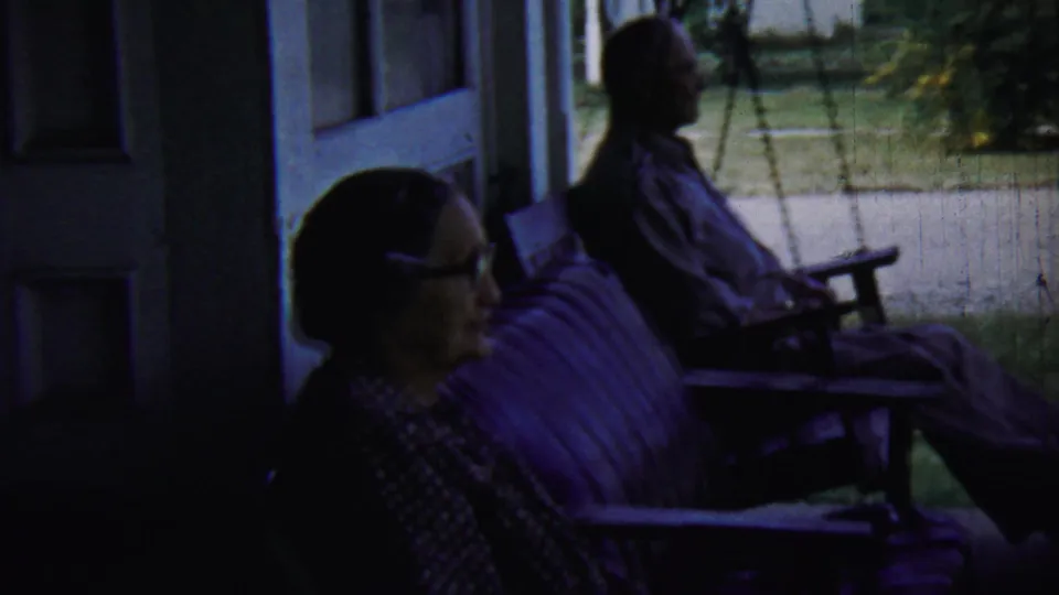 The 1959 home-movie frame shows the seated pair in profile on a porch swing, with yard and walkway visible beyond the shade.