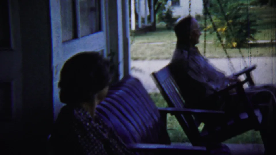 The clip ends with the older adults still seated on the shaded porch swing, the residential yard and neighboring houses beyond.