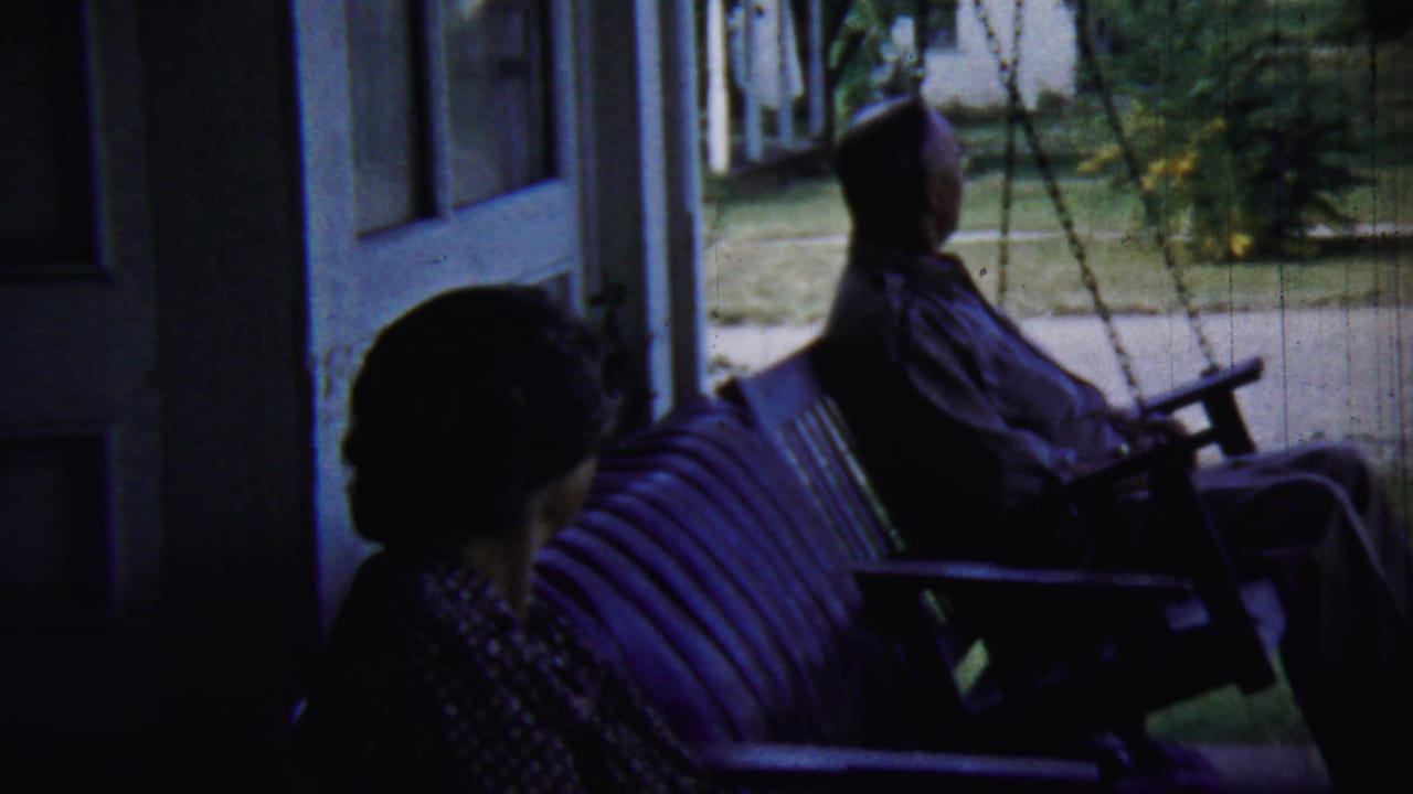 Two older adults sit on a shaded porch, one on a swing and one nearby on a bench.
