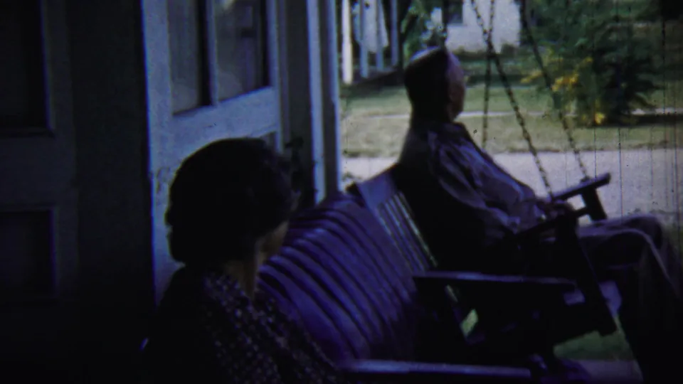 In 1959 Chadron, Nebraska home-movie footage, two older adults sit in silhouette on shaded porch furniture facing a bright yard.