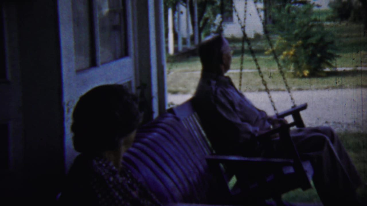 The porch scene continues with the older man on the swing and the older woman beside him, both backlit by the summer yard.