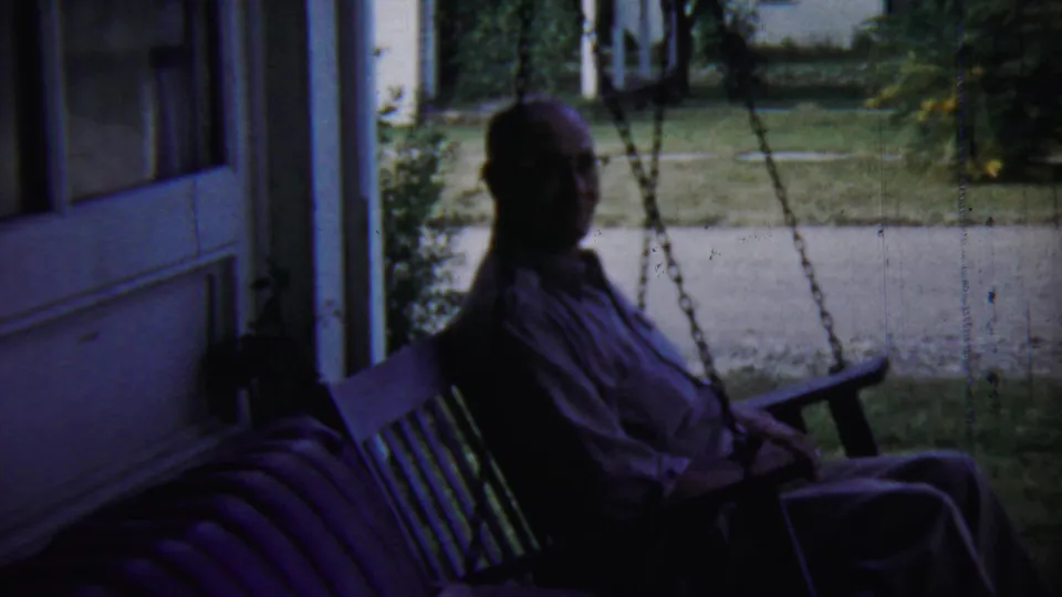 The older man remains seated on the porch swing and appears to glance toward the camera, with the Chadron yard bright behind him.