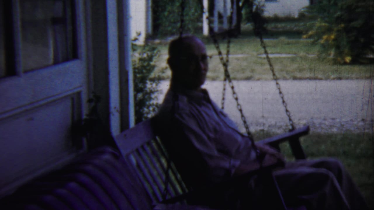 Man seated on a shaded porch swing beside a house doorway with lawn beyond.