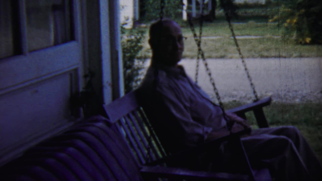Man on a wooden porch swing turns slightly toward the camera in porch shade.