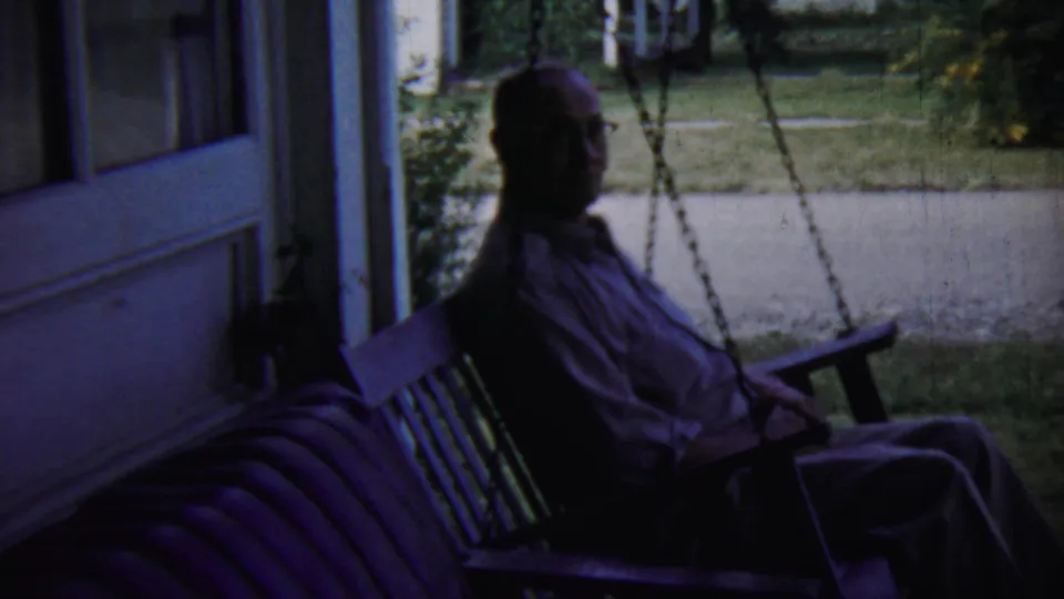 The seated man remains on the porch swing, partly silhouetted against the brighter yard in the 1959 Chadron home-movie footage.