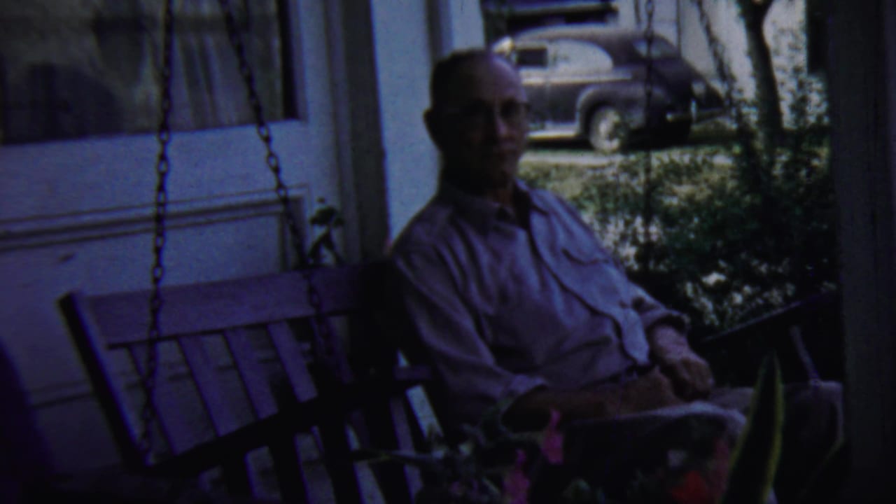 Man seated on a porch swing with a parked vintage car visible beyond the porch.