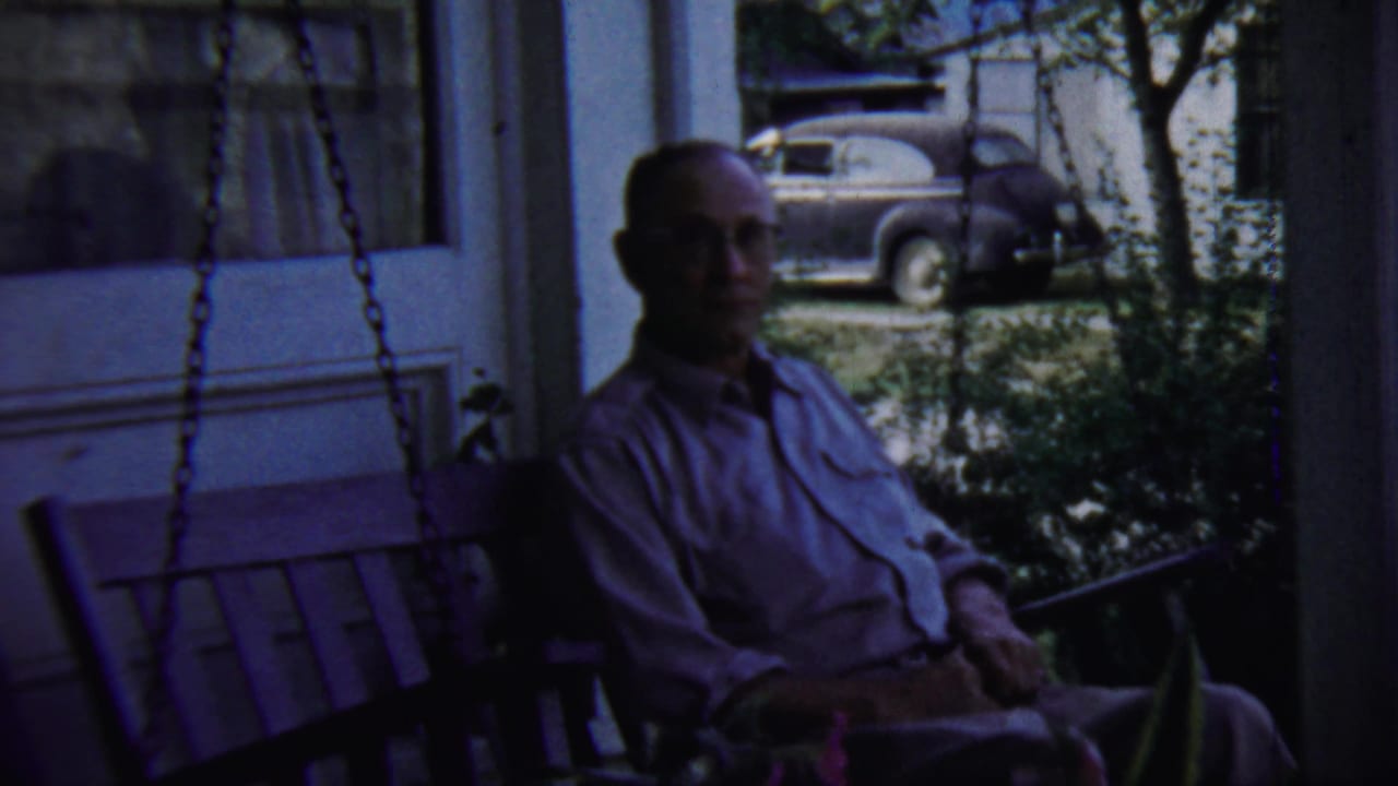 The 1959 Chadron, Nebraska frame shows the man seated on the porch swing, partly shaded, with a rounded mid-century sedan outside.
