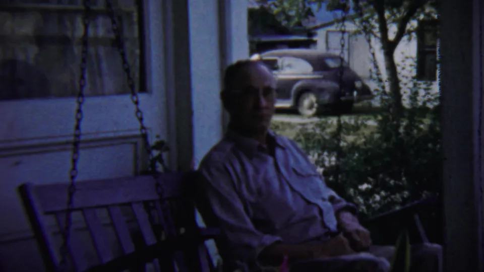 A man in a light shirt remains seated on a porch swing in the 1959 Chadron home-movie scene, with greenery and a vintage car behind him.