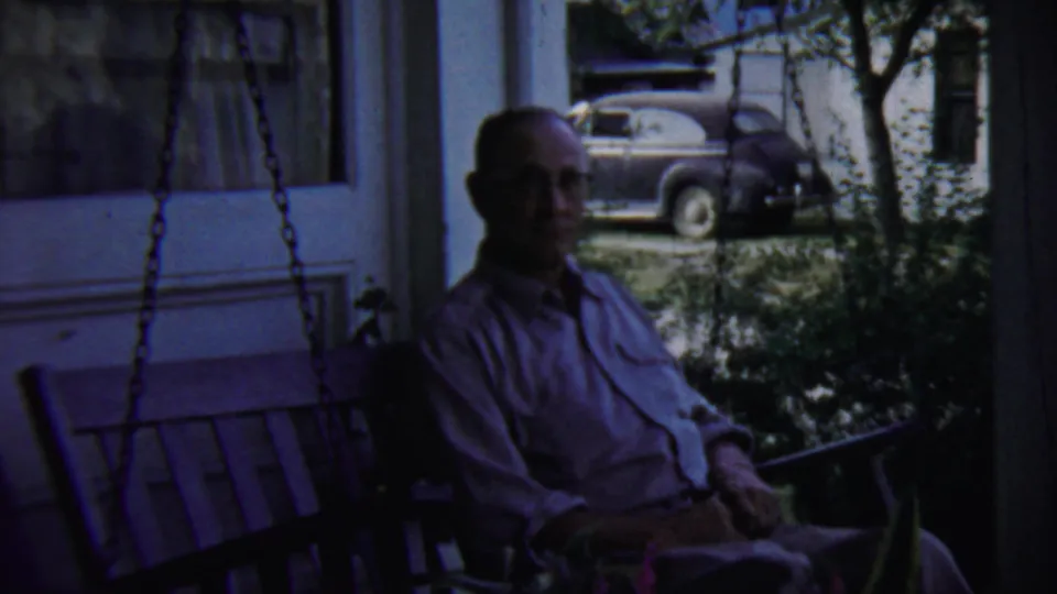 Final 1959 Chadron, Nebraska frame of the man seated on the shaded porch swing, with a house exterior, plants, and 1950s car beyond.