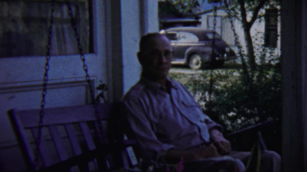 Man with glasses sitting on a porch swing with a parked car visible outside.
