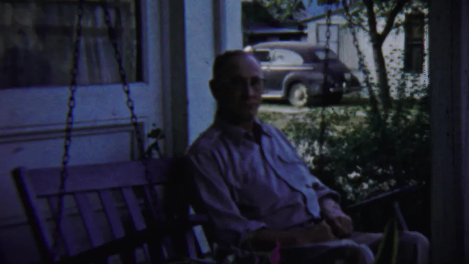 In Chadron, Nebraska home-movie footage, a man sits on a shaded porch swing while a vintage car is visible beyond the porch.