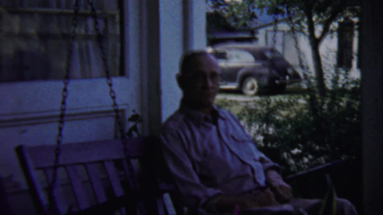A seated man remains on the porch swing in 1959 Chadron footage, framed by porch posts, foliage, and a parked period car.