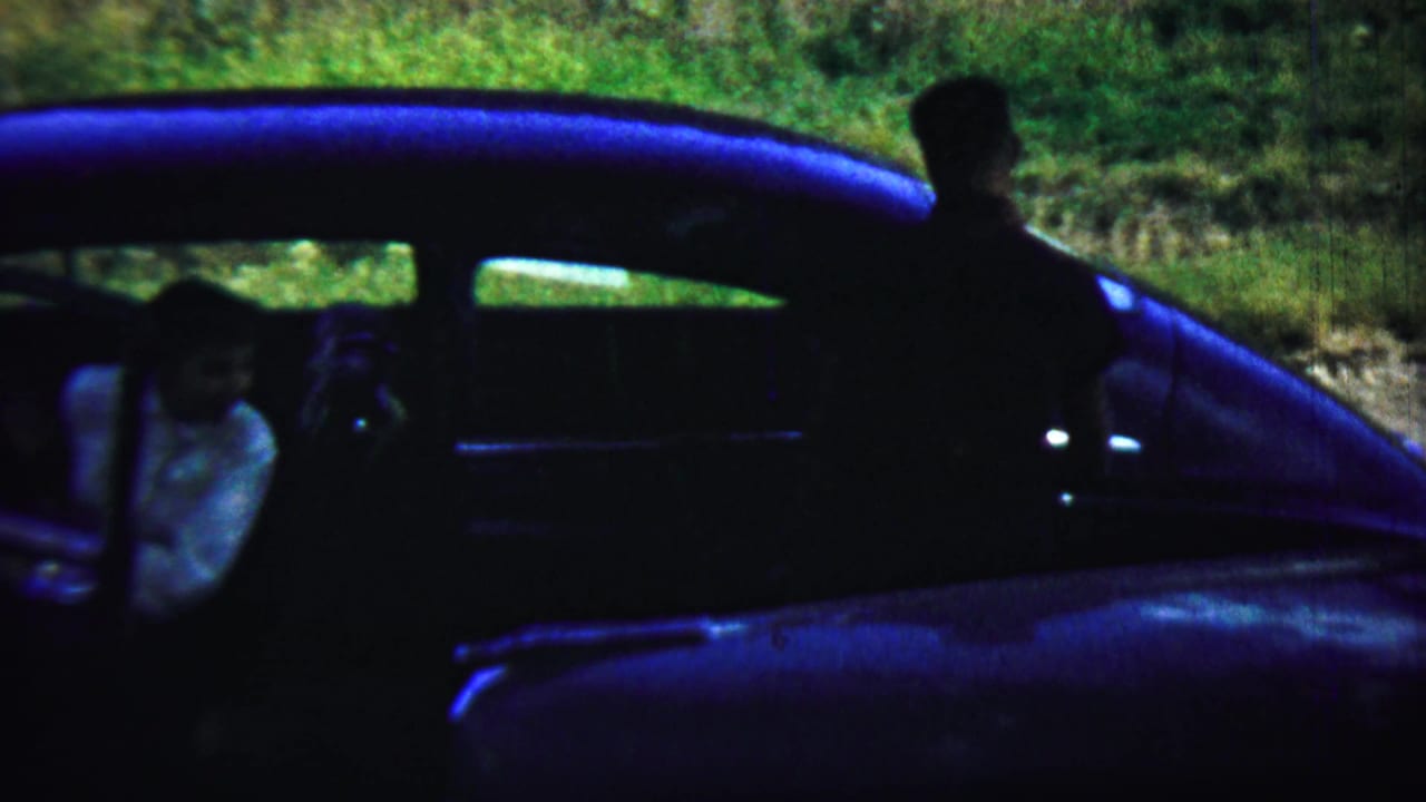 Two adults beside a dark blue car on a sunlit gravel surface.