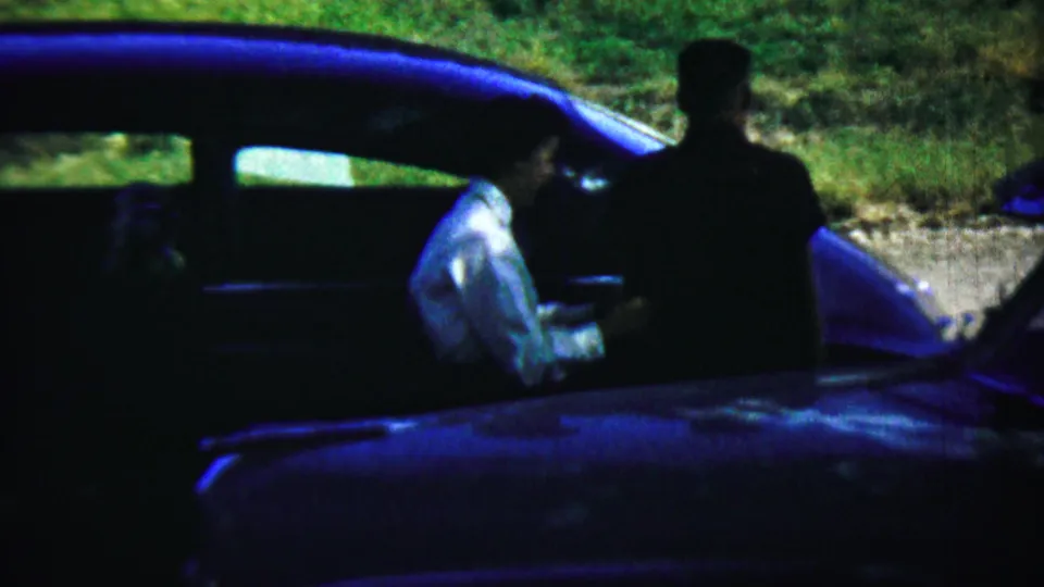 A light-shirted adult stands beside a blue car in Chadron, Nebraska, with another dark-clothed figure near the vehicle.