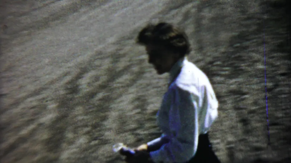 A woman in a white blouse and dark skirt walks away from the blue car across a gravel surface in the 1959 Chadron home movie.
