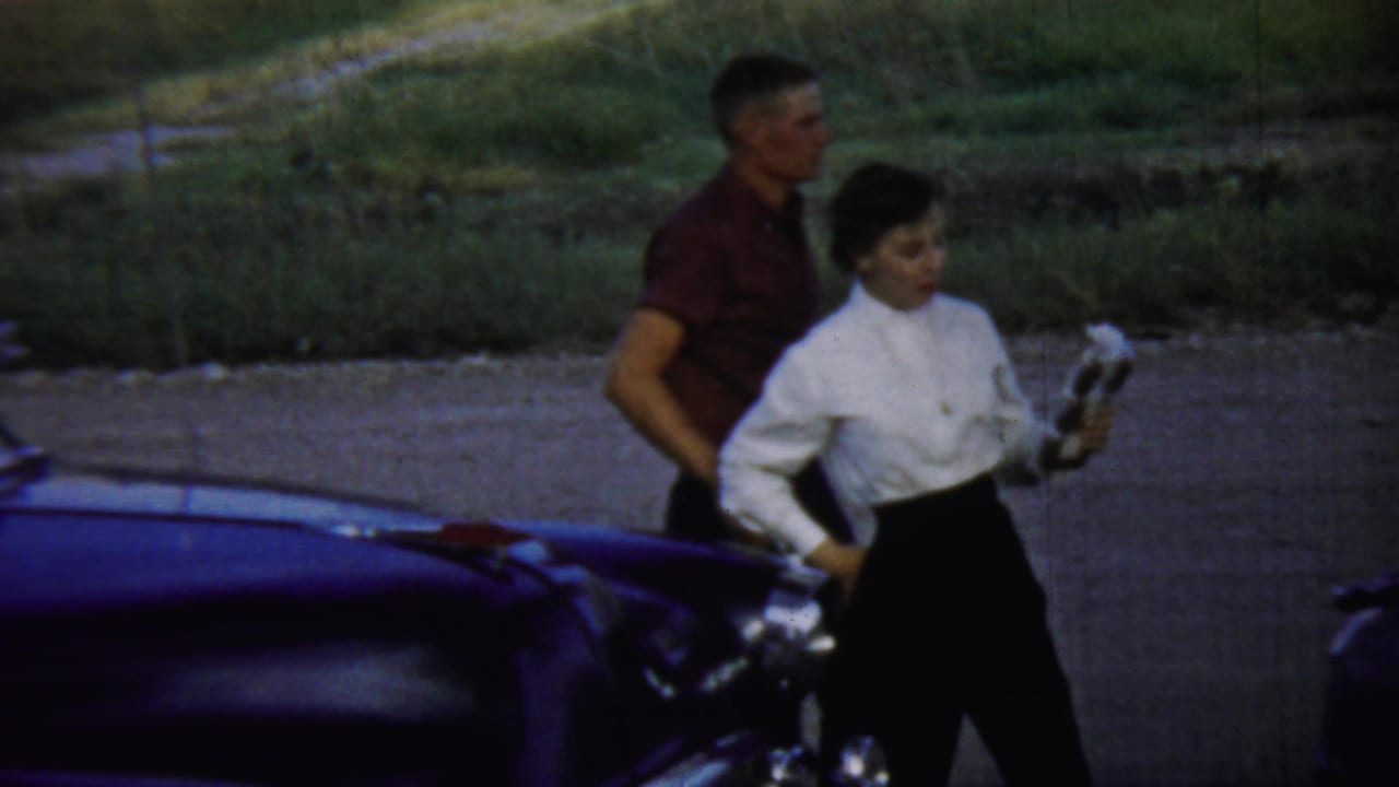 Young couple walking beside a dark classic car on gravel near grass.