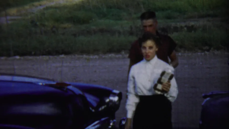 In the Chadron, Nebraska home-movie scene, the woman walks toward camera holding a small object while the man follows near the car.