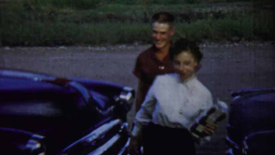 The couple smiles while walking past the rounded front end of a dark classic car in the 1959 Chadron, Nebraska footage.
