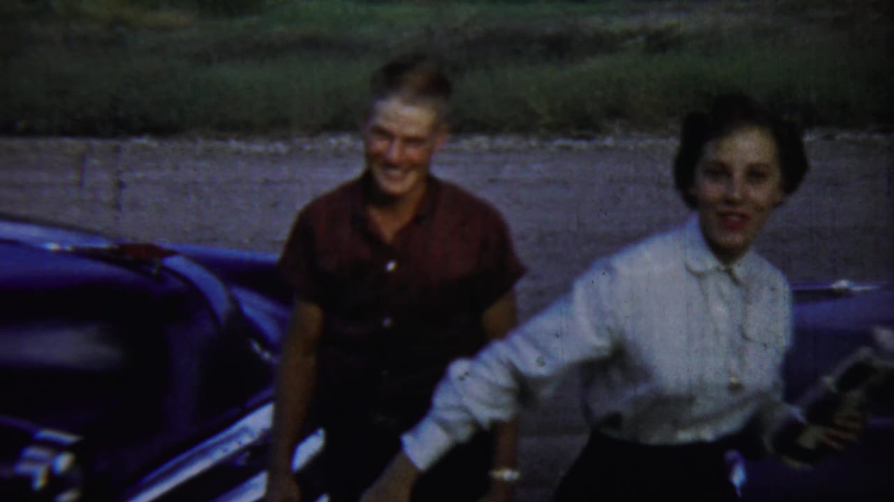 Smiling man and woman standing beside a dark classic car outdoors.