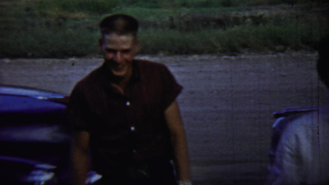 Man in a maroon shirt smiles near a dark classic car.