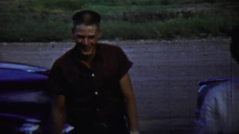 A man in a maroon short-sleeve shirt smiles near a dark classic car in 1959 Chadron, Nebraska home-movie footage.