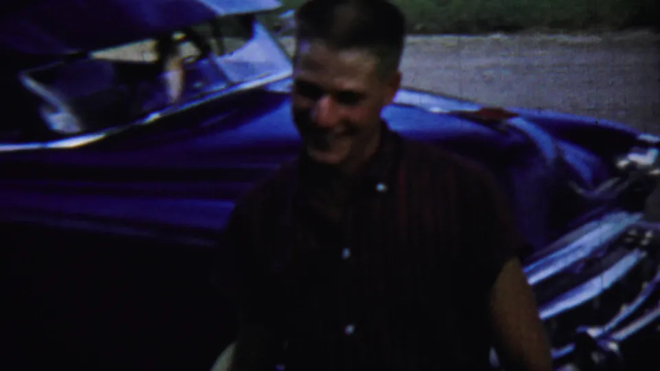 A smiling man stands close to a dark classic car, with deep shadow and saturated 8mm color in the frame.