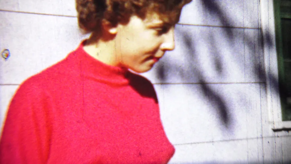A woman in a red turtleneck is shown in profile near a light exterior wall, likely from the same 1959 Chadron home-movie reel.