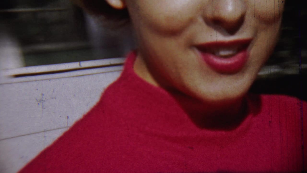 Close handheld view of the woman smiling in a red turtleneck, with a wall and window behind her in the 1959 Chadron home movie.