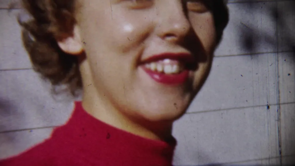 Smiling young woman in a red turtleneck fills the frame, with pale wall panels behind her in 1959 Chadron home-movie footage.
