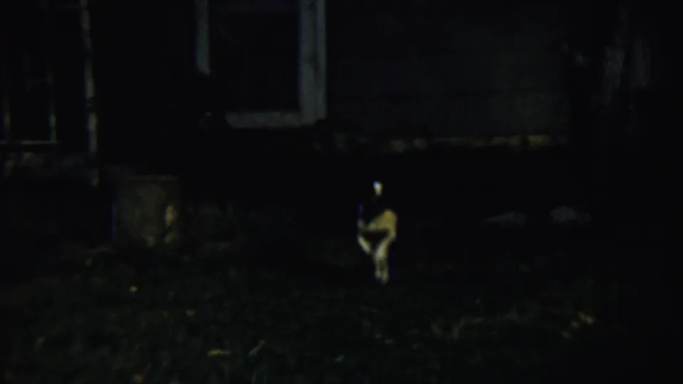1959 Chadron, Nebraska 8mm frame showing a small dog in a dark grassy yard near a dim building exterior, lit by the camera.