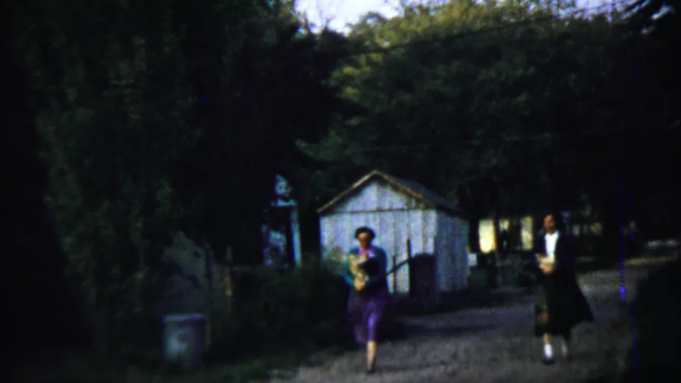 Two women walk along a path near trees and a small shed in Chadron, Nebraska, at the end of the 1959 home movie clip.