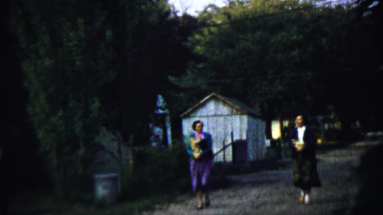 Two traditionally dressed adults walk on a gravel path near trees and a white shed.
