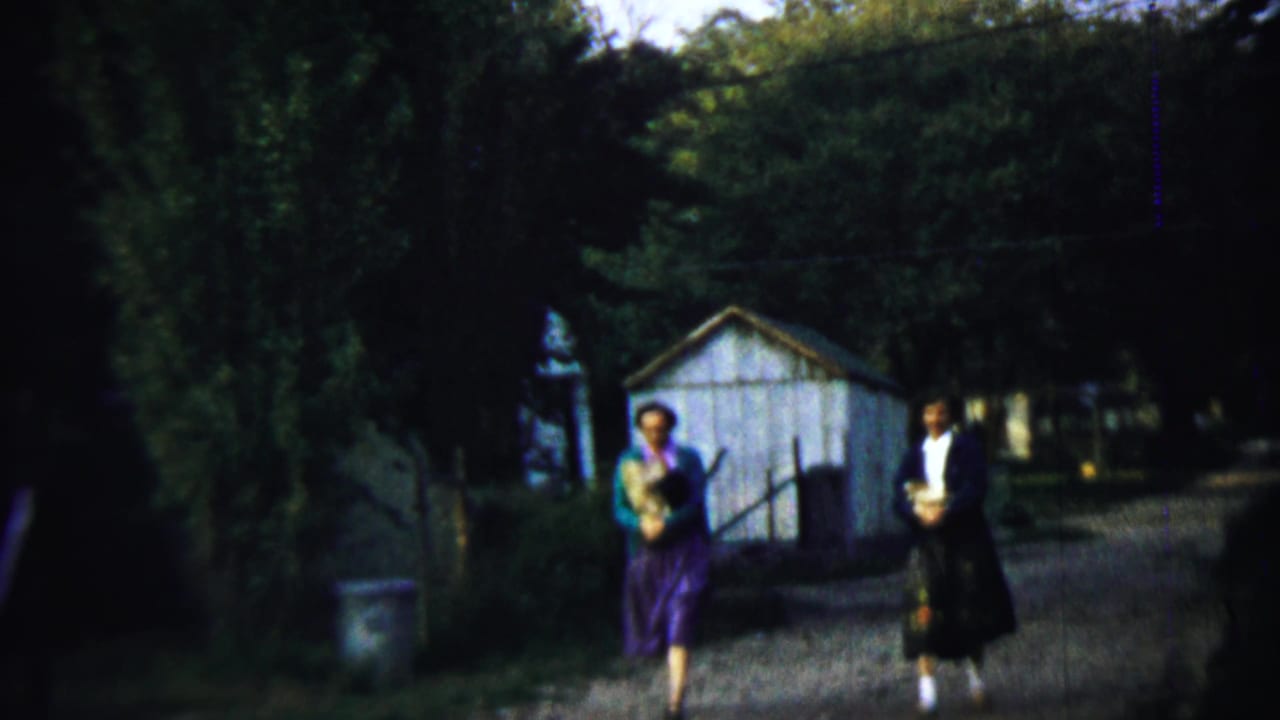 The two adults continue along the rural path in Chadron, Nebraska, with bundles in hand and a small white shed behind them.