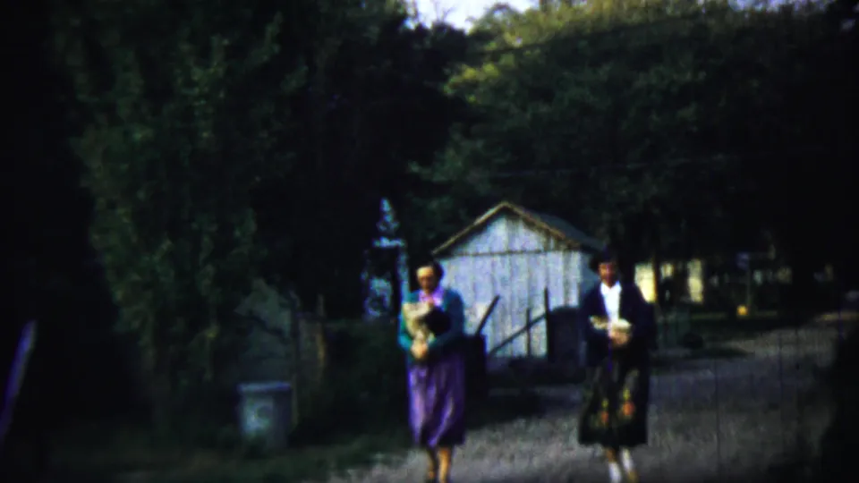 Two traditionally dressed adults walk closer to the camera on a shaded gravel path in the 1959 Chadron, Nebraska home-movie scene.