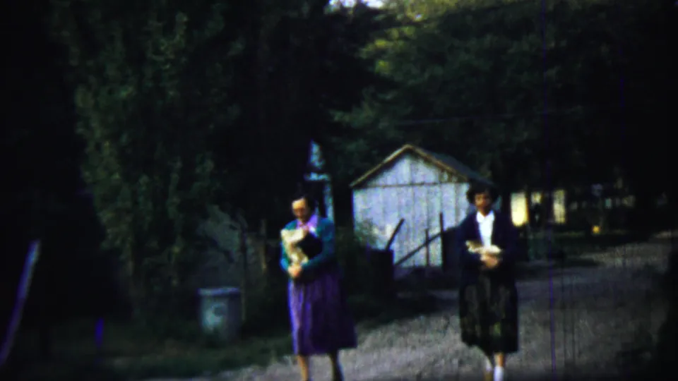 Near the end of the Chadron, Nebraska clip, the two adults pass closer to camera with bundles, trees, fencing, and the white shed in view.