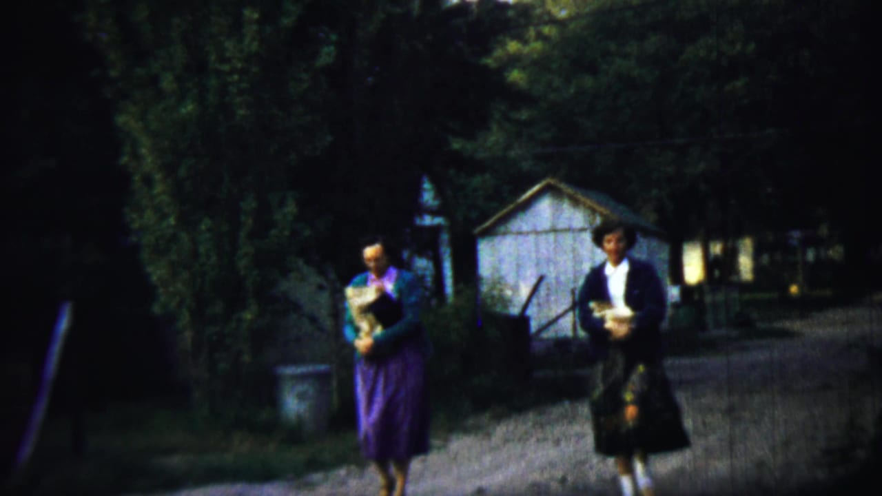 Two women walk on a dirt path near trees, a small shed, and a house.
